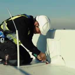 Pracownik w kasku i uprzęży zabezpieczającej, uszczelnia połączenie na białym dachu płaskim przy użyciu pistoletu do uszczelniacza.
