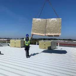 Pracownik w kasku i uprzęży asekuracyjnej kieruje dźwigiem z paletą wełny mineralnej na dachu z blachy trapezowej w Przemyślu, przygotowując się do montażu.