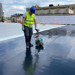 Pracownik w kasku i kamizelce odblaskowej zgrzewa czarną membranę dachową na płaskim dachu, w tle budynki miejskie i częściowo zrujnowana kamienica z dekoracyjnym rysunkiem elewacji.