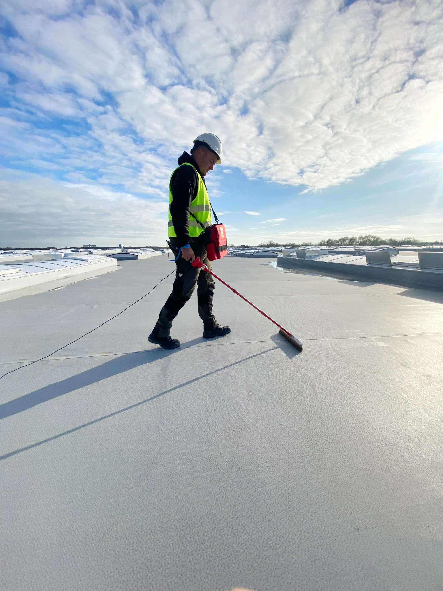 Pracownik w kasku i kamizelce odblaskowej rozprowadza płynną substancję po powierzchni dachu za pomocą szczotki na długim kiju, na tle błękitnego nieba z chmurami i świetlików dachowych.