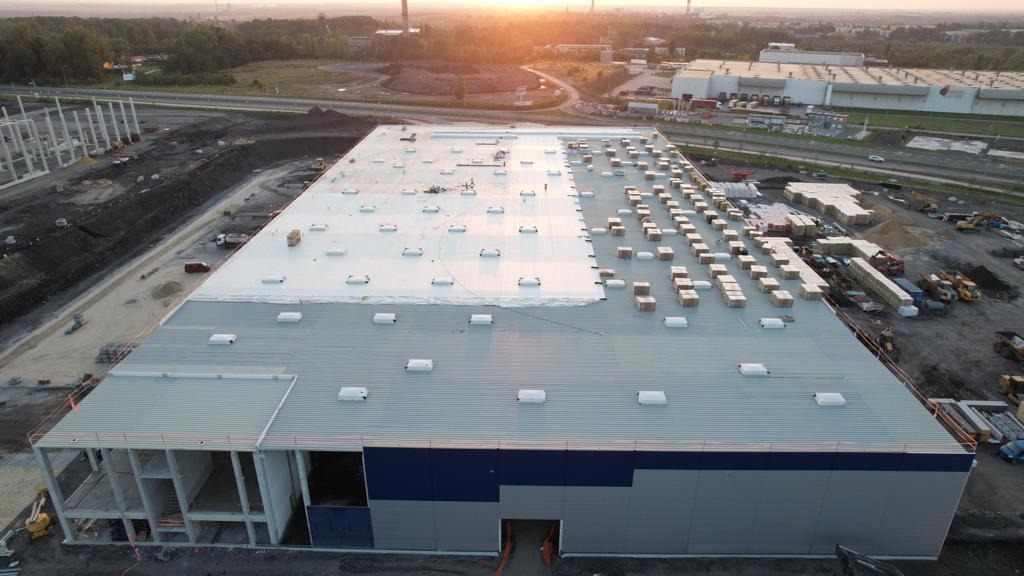 Szeroki kadr z lotu ptaka ukazuje budynek przemysłowy z częściowo wykonanym jasnym pokryciem dachowym; materiały budowlane ułożone w rzędach na dachu, w tle widoczne inne budynki przemysłowe...