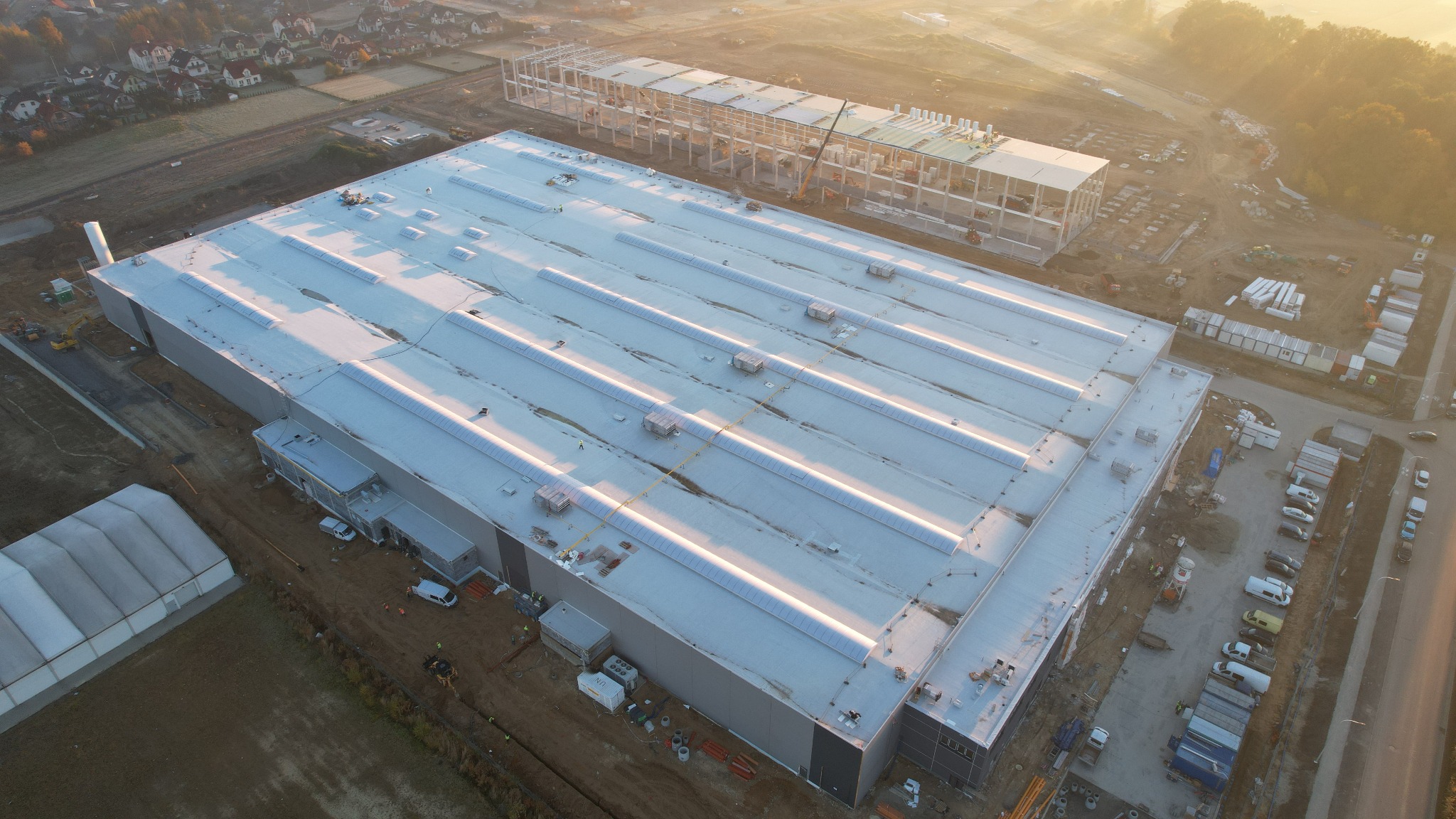 Rozległy dach hali przemysłowej z świetlikami, widziany z lotu ptaka w porannym słońcu, obok wznoszona konstrukcja stalowa, teren budowy z kontenerami i pojazdami.