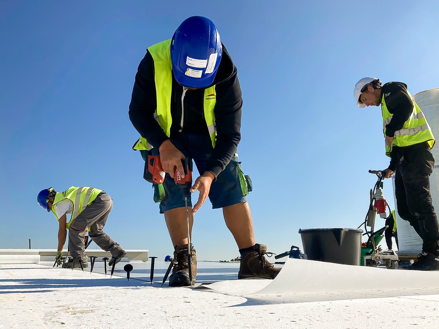 Montaż izolacji dachu płaskiego przy użyciu wkrętarki akumulatorowej Hilti i łączników mechanicznych; trzech pracowników w odblaskowych kamizelkach i kaskach na tle błękitnego nieba.