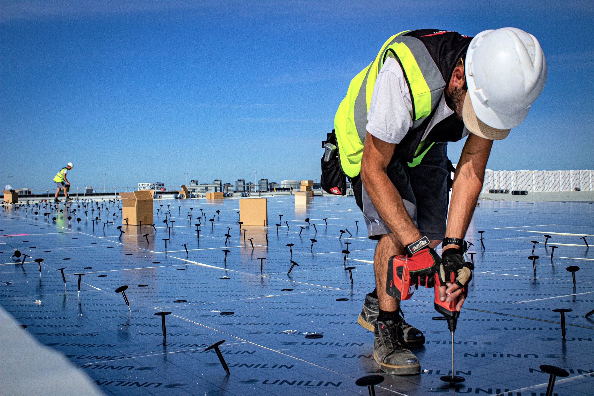 Pracownik w kasku i kamizelce odblaskowej montuje elementy mocujące na folii dachowej Unilin, widoczne kartony i inni pracownicy w tle.