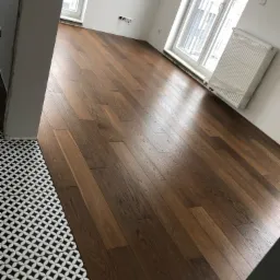 Ukończona podłoga z ciemnego drewna ułożona w jodełkę, sąsiadująca z czarno-białą mozaiką w geometryczny wz&oacute;r przy progu drzwi, widok na białe ściany, okno i grzejnik.