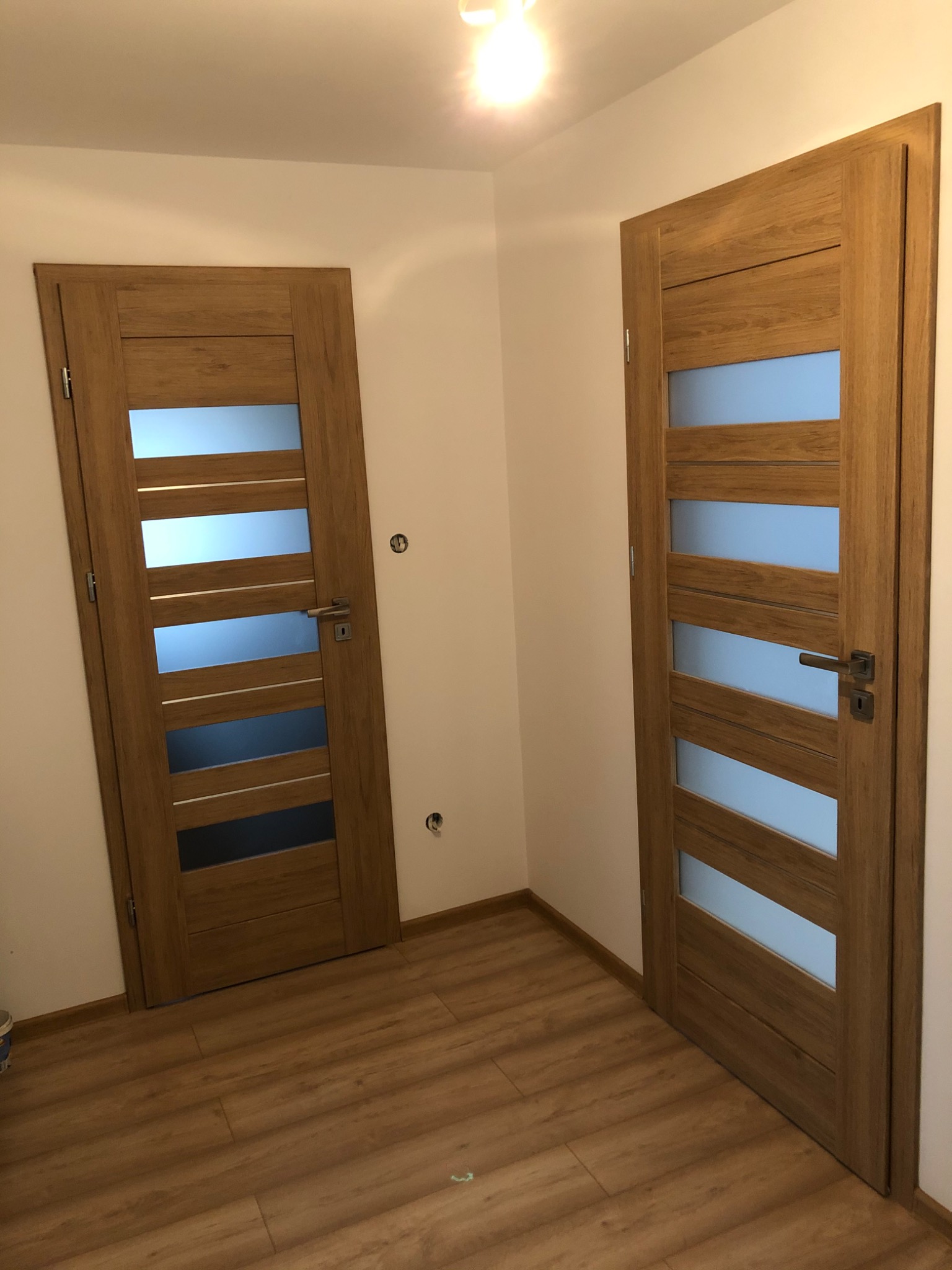 Dwie drewniane drzwi wewnętrzne z przeszkleniami, zamontowane w jasnym pomieszczeniu z drewnopodobną podłogą, widoczne ślady po otworach montażowych na ścianie.