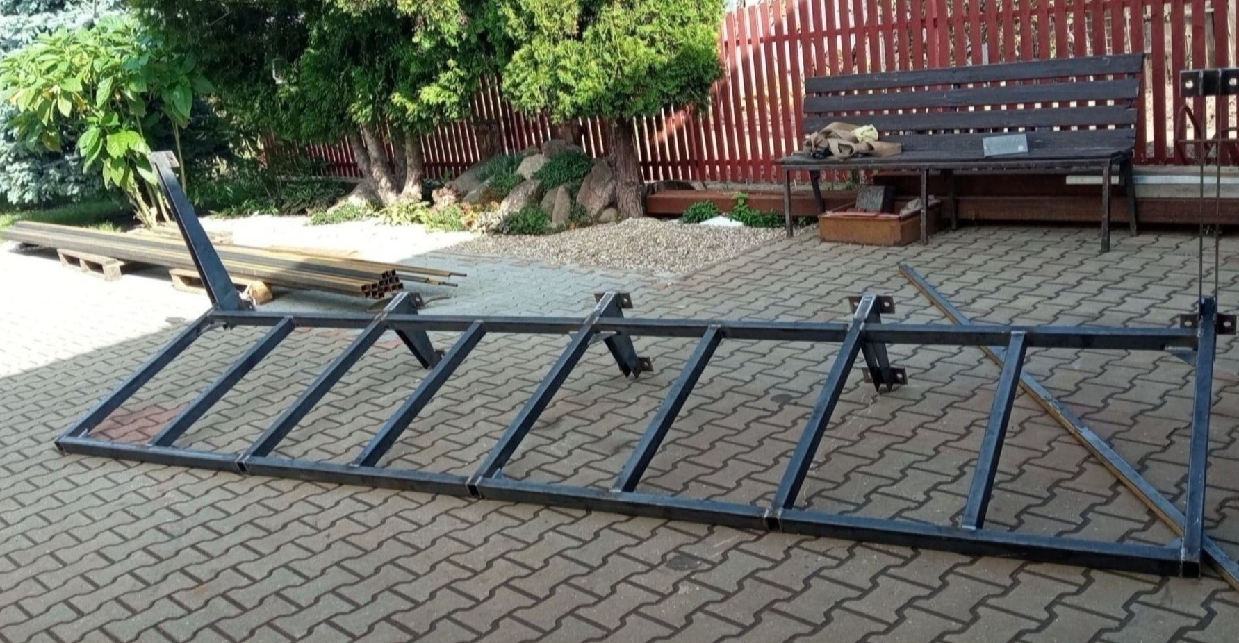 Metalowa konstrukcja ramy bramy w trakcie budowy, leżąca na brukowanej powierzchni, z widocznymi spawami i elementami mocującymi, w tle ogród z czerwonym płotem.