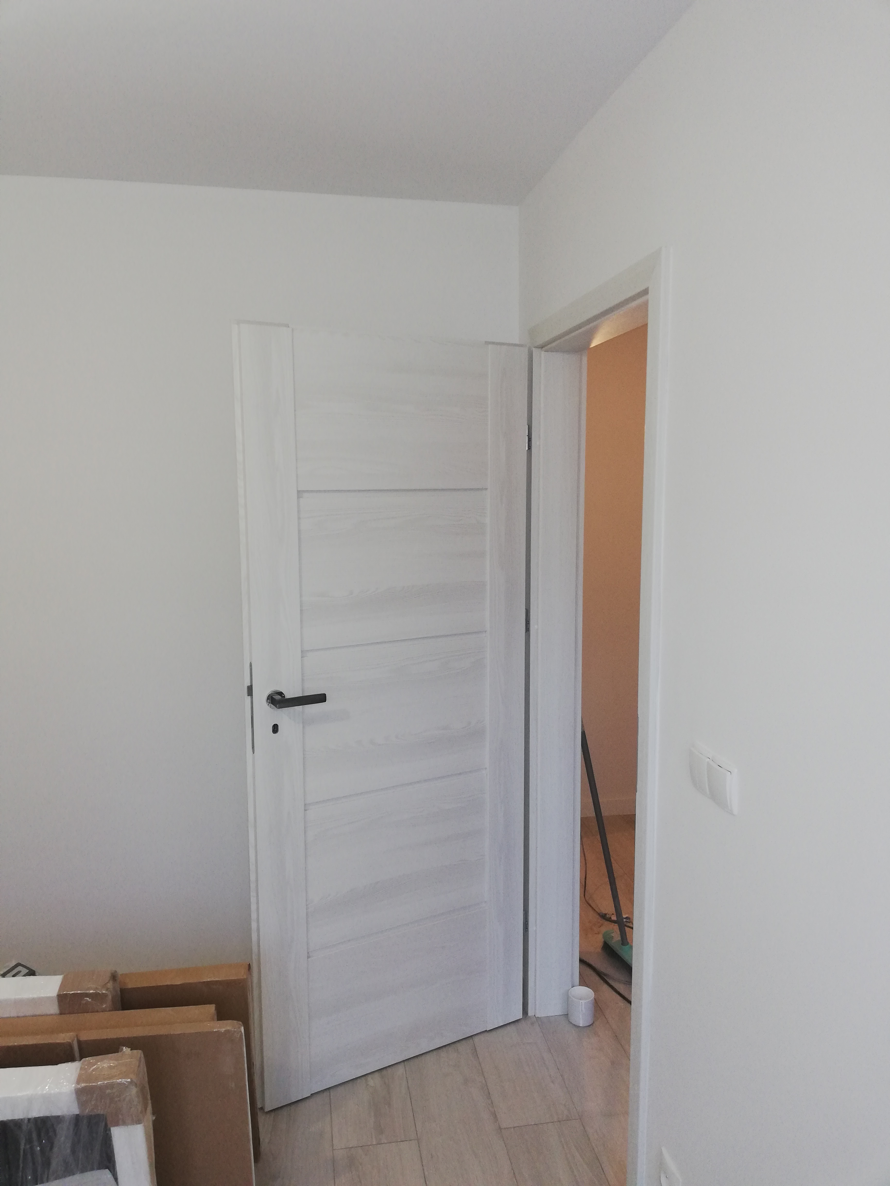 Montaż wewnętrznych drzwi drewnianych z czarną klamką w nowym mieszkaniu, widoczne kartony po drzwiach i listwa przypodłogowa w tle.