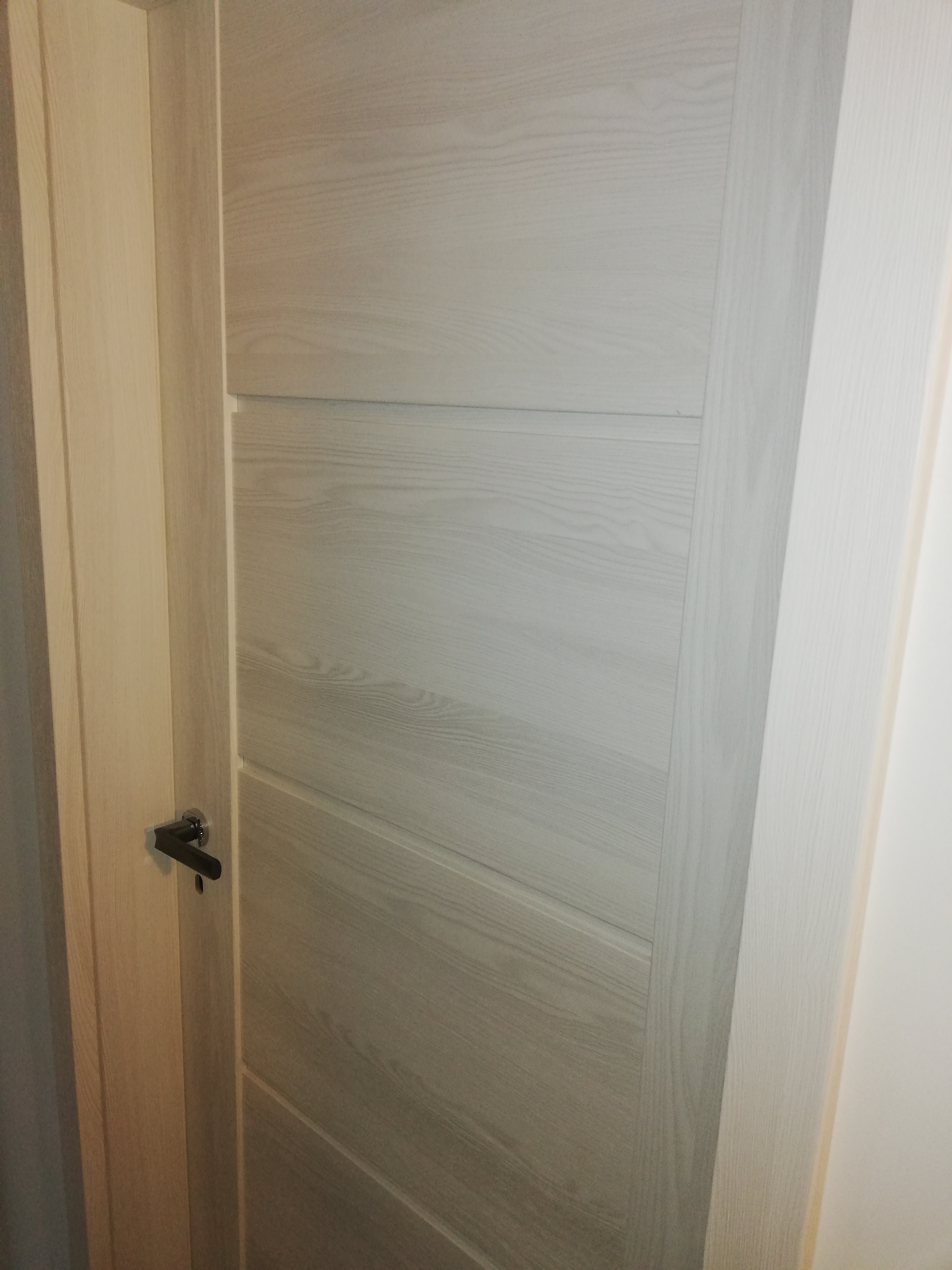 Zamknięte, drewniane drzwi wewnętrzne o jasnym odcieniu z poziomymi panelami i czarną klamką.