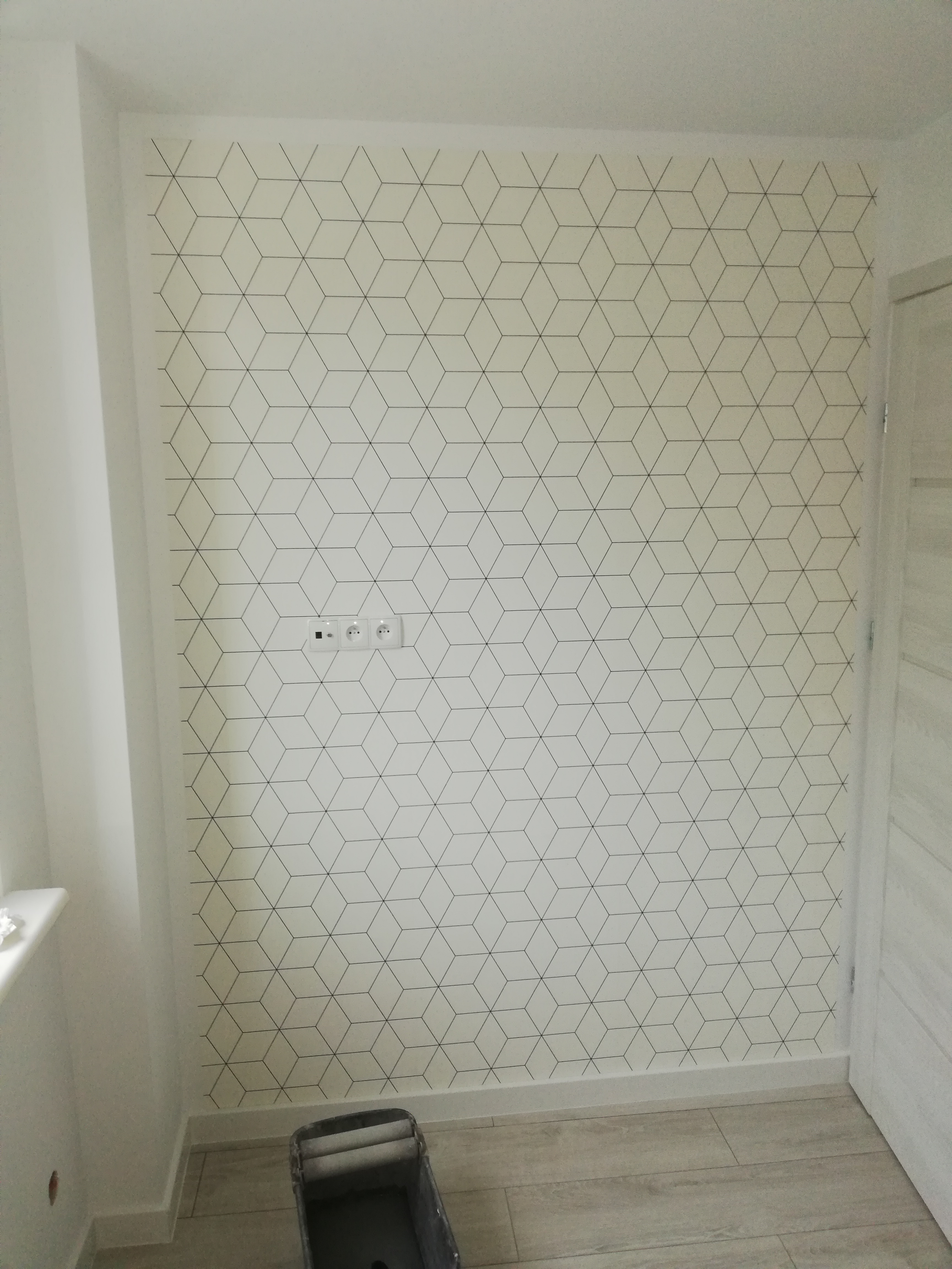 Ściana po remoncie z białą tapetą w geometryczny wzór czarnych sześcianów, widoczne gniazdka elektryczne i wiaderko malarskie przy podłodze.
