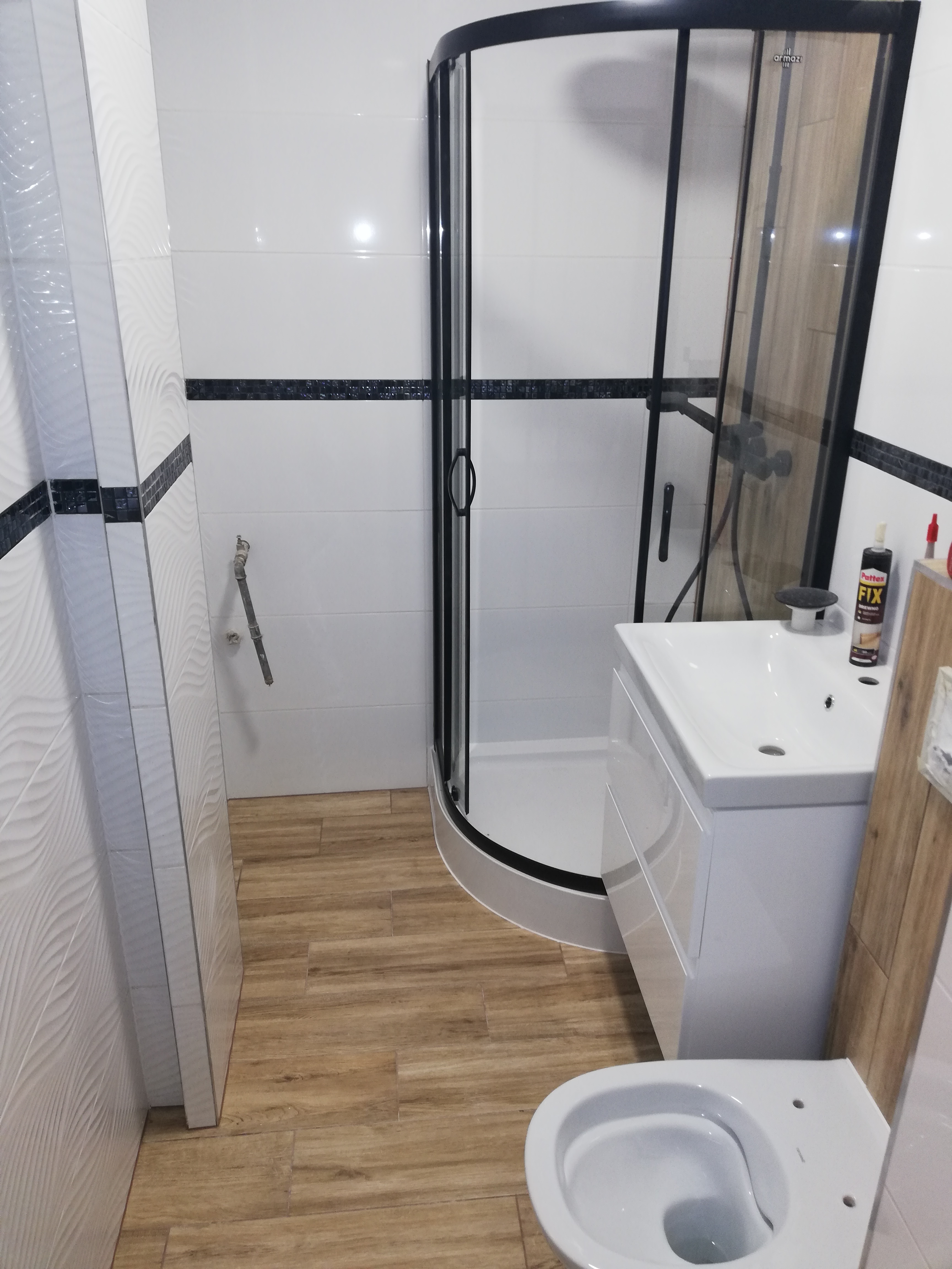 Wykończona łazienka z kabiną prysznicową z czarnymi ramami, białą umywalką z szafką, toaletą i płytkami imitującymi drewno na podłodze. Na umywalce stoi klej Pattex FIX.