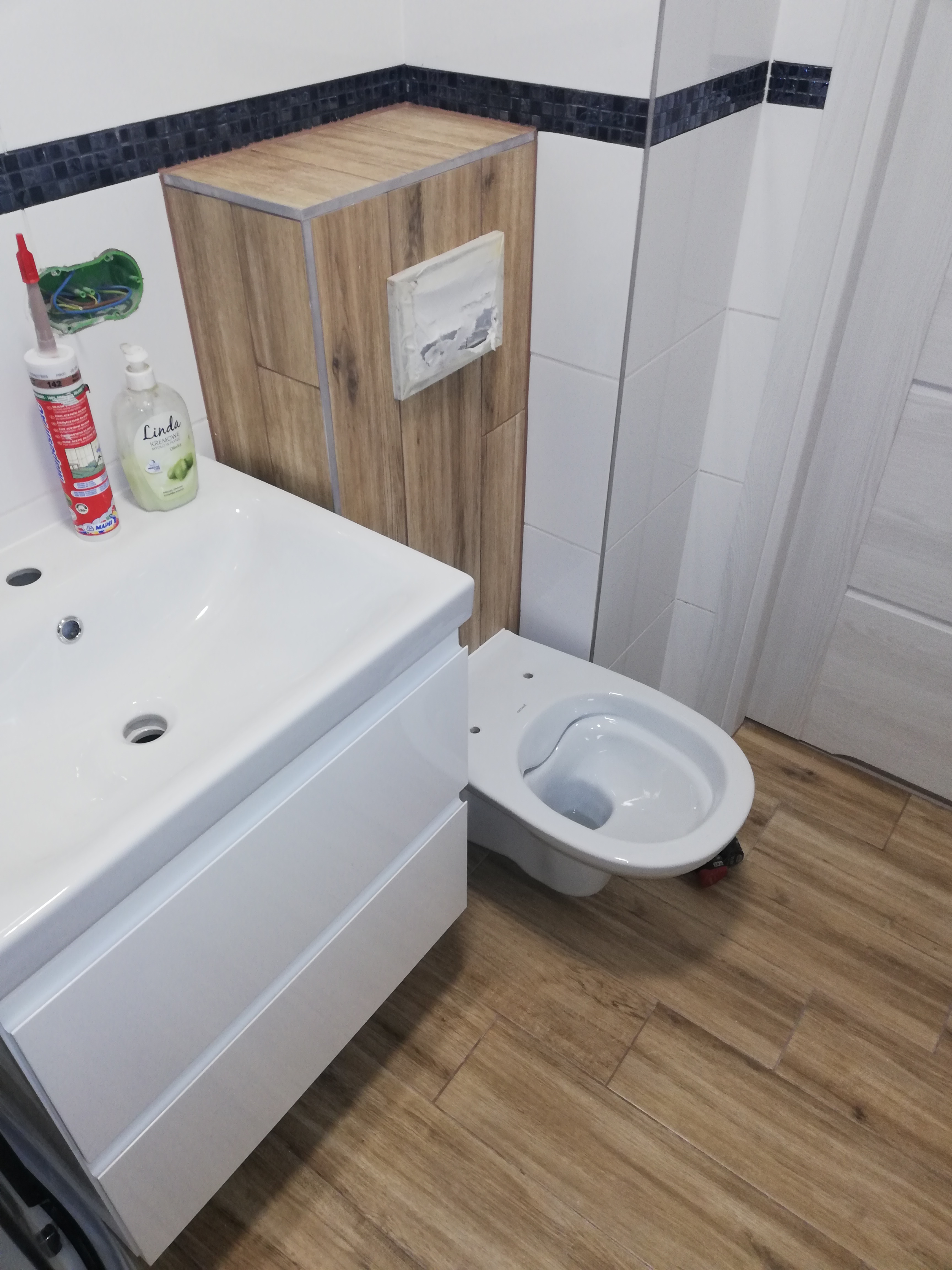 Umywalka z szafką, wisząca toaleta i zabudowa stelaża WC z płytek imitujących drewno w łazience po remoncie, widoczne narzędzia i materiały budowlane.