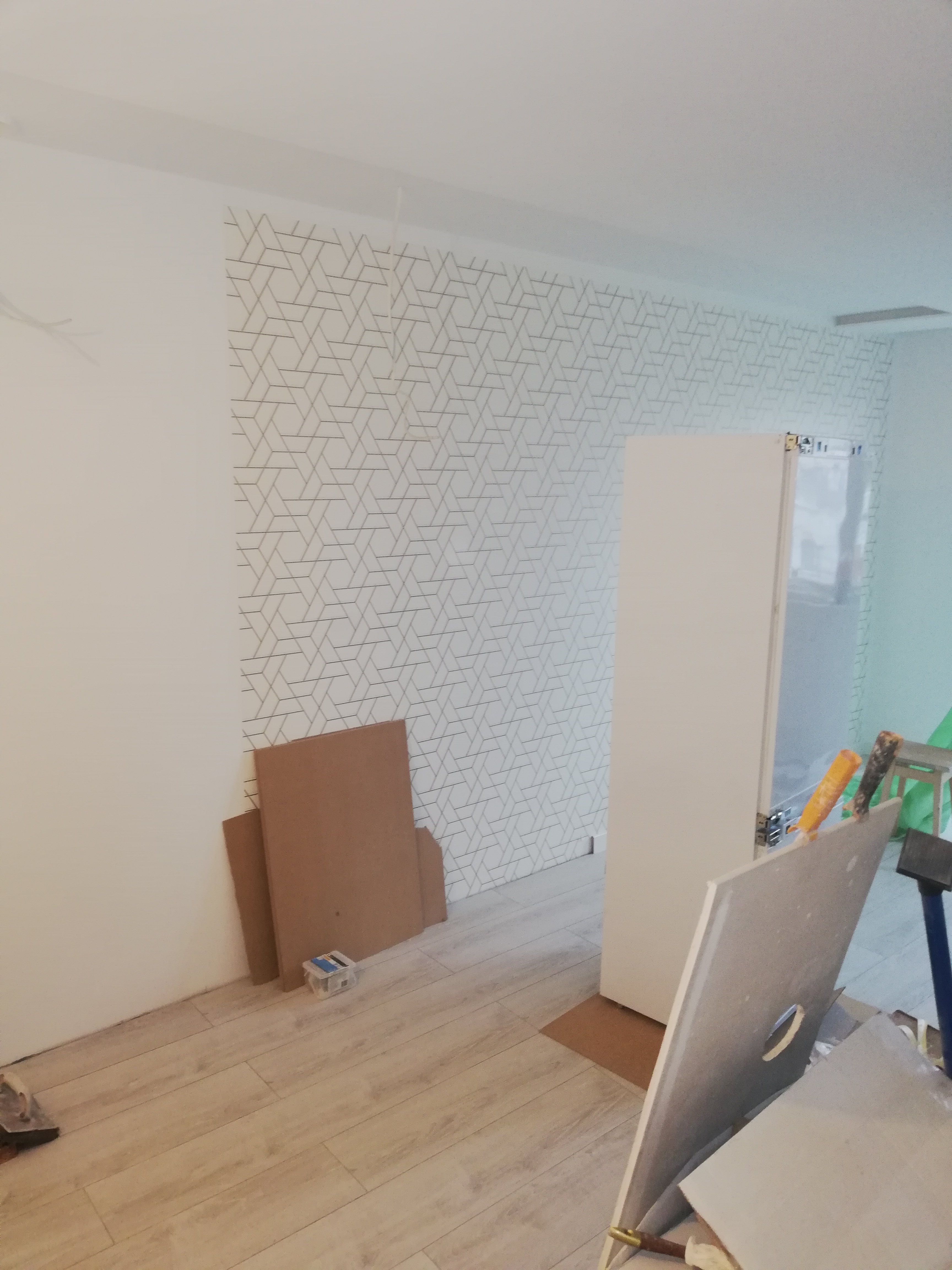 Ściana z geometryczną tapetą w trakcie remontu, widoczne płyty gipsowe, kartony i narzędzia w pomieszczeniu z jasną podłogą.