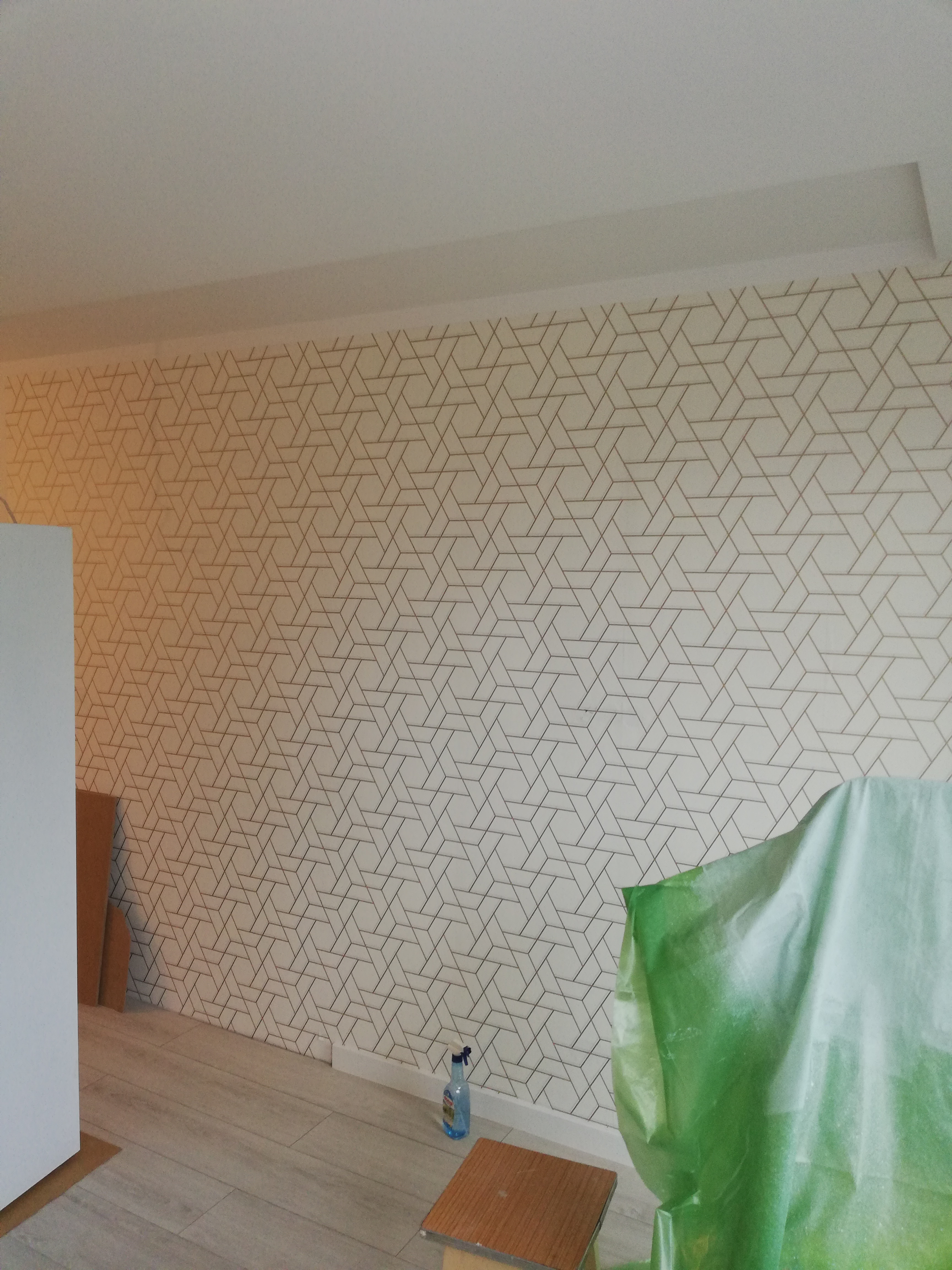 Ściana z geometryczną tapetą w trakcie zabezpieczania folią malarską, widoczny spryskiwacz i stołek.
