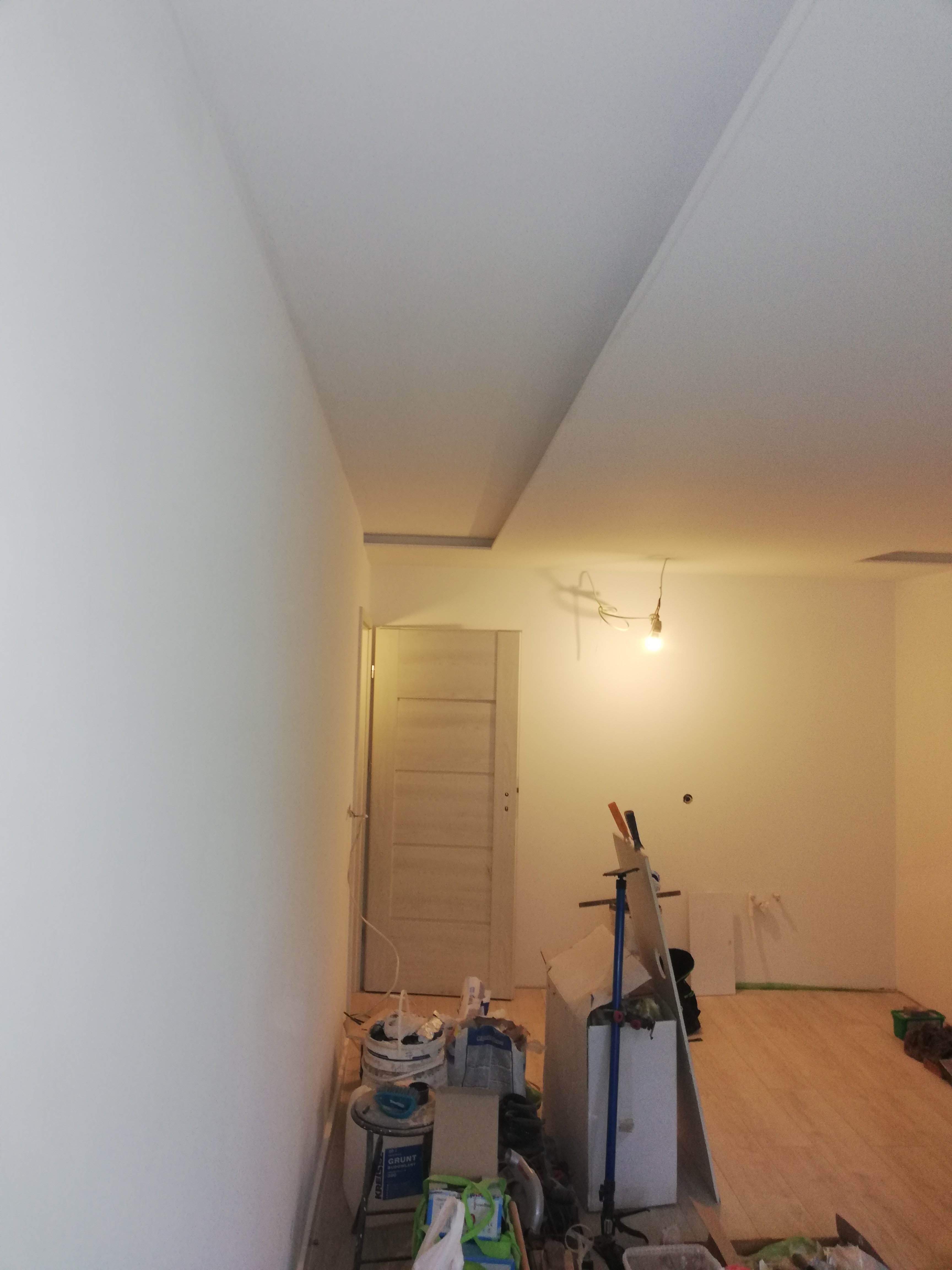 Wnętrze w trakcie malowania ścian na biało, widoczne narzędzia malarskie, drabina, wiadro z farbą oraz niezabezpieczona podłoga z paneli.