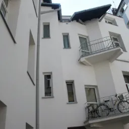 Biała elewacja budynku z balkonami, na jednym z nich stoi rower, widok z dołu.