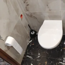 Nowoczesna toaleta wisząca z przyciskiem spłukującym Cersanit, czarną szczotką do WC i uchwytem na papier toaletowy, wykończona marmurowymi płytkami na ścianach i podłodze.