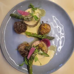 Dwa talerze z elegancko podanym daniem: roladki z mięsa, pieczone mini-ziemniaki w harmonijkę, szparagi, marynowana cebulka i sos, udekorowane świeżymi ziołami.