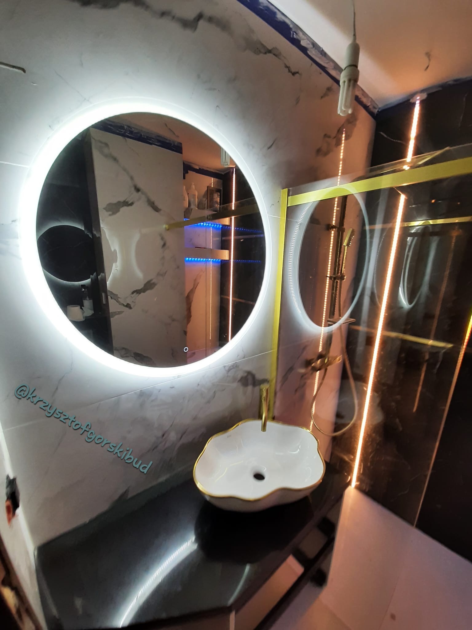 Nowoczesna łazienka z okrągłym lustrem LED, umywalką w kształcie kwiatu z złotym obramowaniem i kabiną prysznicową z złotymi akcentami na tle marmurowych płytek.