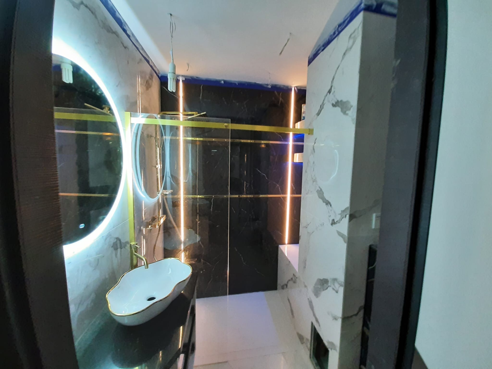 Nowoczesna łazienka w trakcie wykańczania: czarne marmurowe ściany z podświetleniem LED, umywalka nablatowa z złotym obramowaniem, kabina prysznicowa z złotymi elementami, białe marmurowe płytki...