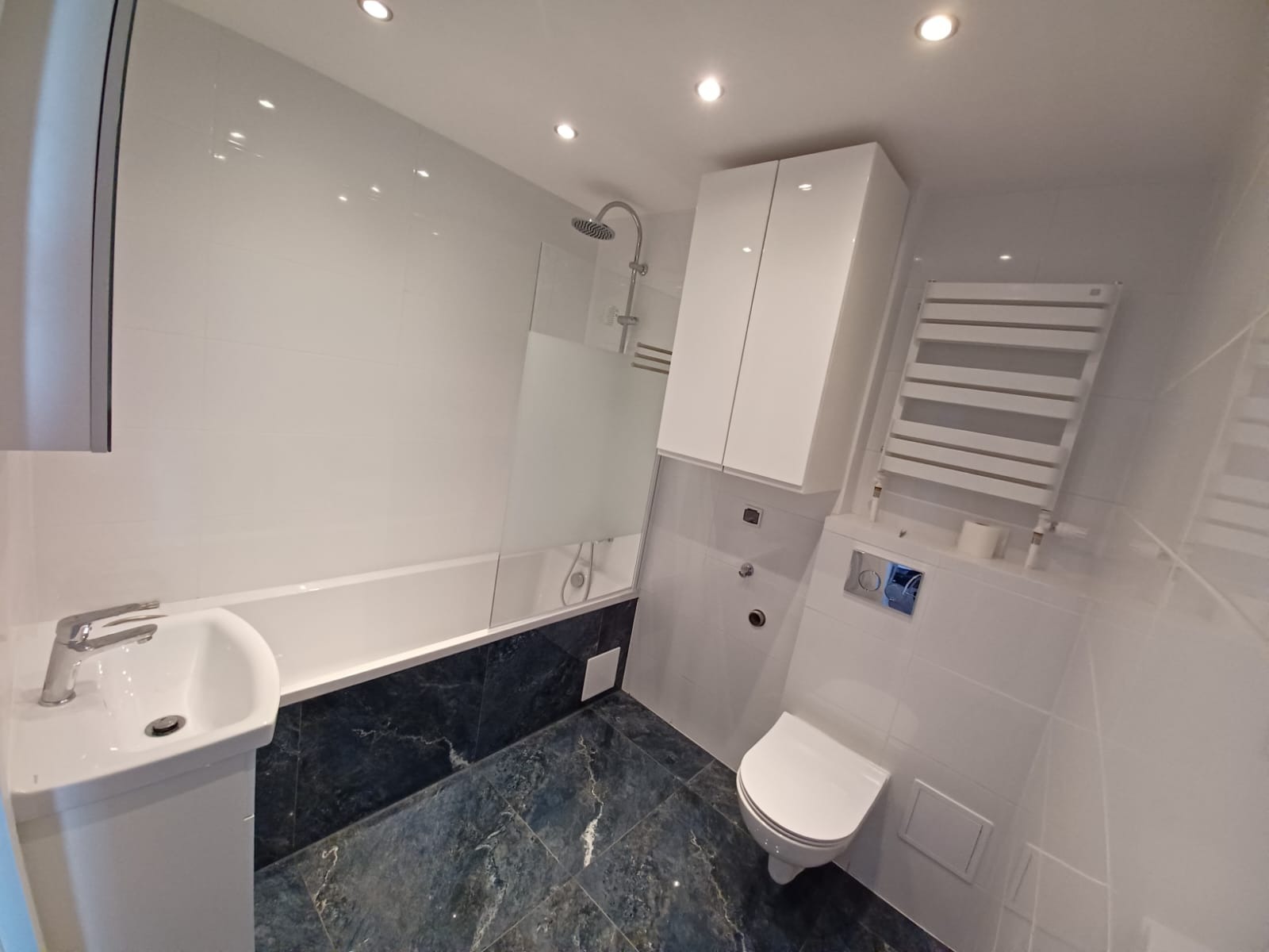 Nowoczesna łazienka z białą armaturą, kabiną prysznicową ze szklanym panelem, szafką nad toaletą i podłogą w ciemnym marmurze.