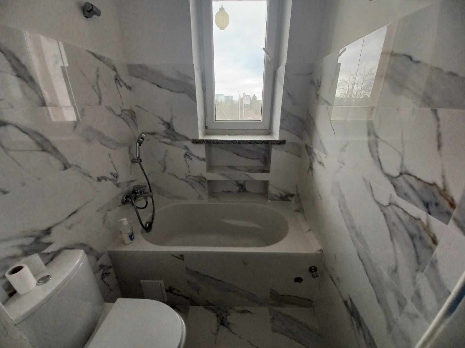 Biała łazienka z wanną, toaletą i płytkami imitującymi marmur, okno w zabudowie z półkami.