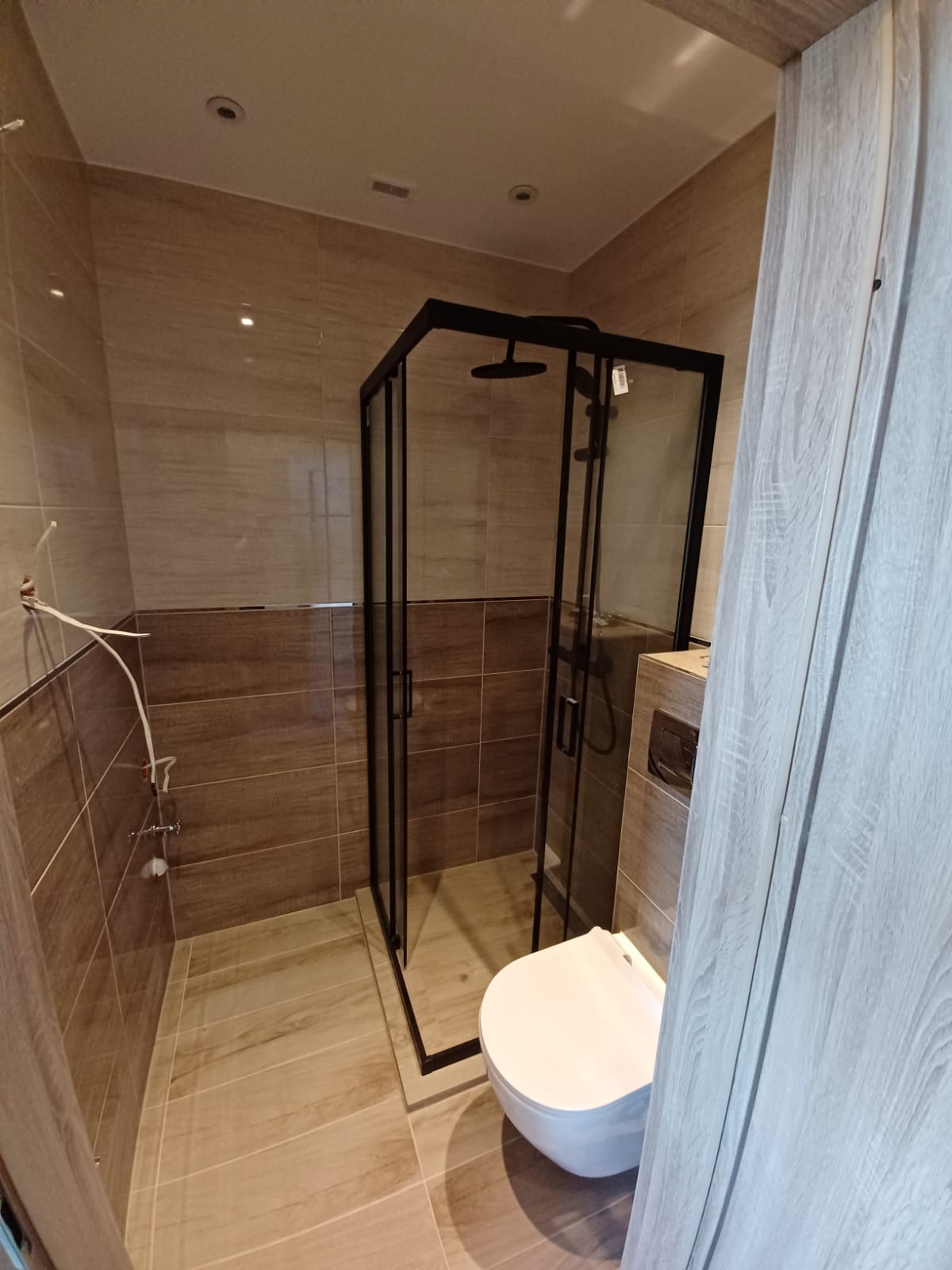 Wykończona łazienka z kabiną prysznicową z czarnymi ramami, wiszącą toaletą i brązowymi płytkami imitującymi drewno.
