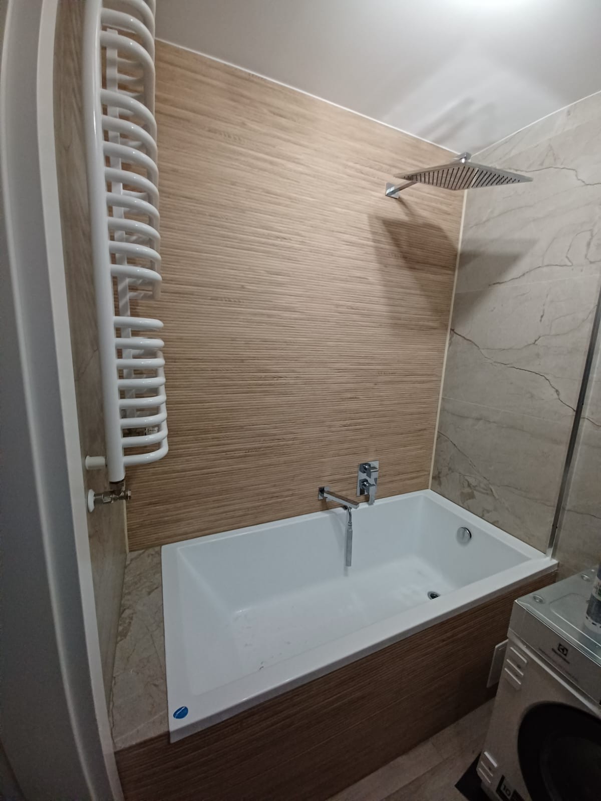Nowoczesna łazienka z białą wanną, baterią ścienną i deszczownicą, wykończona płytkami imitującymi drewno i marmur, z grzejnikiem drabinkowym po lewej stronie i pralką widoczną w prawym dolnym rogu.
