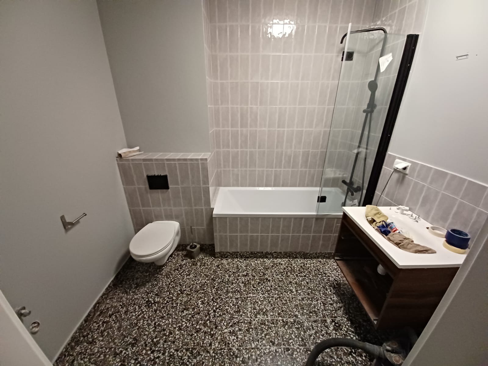 Wyremontowana łazienka z mozaikową podłogą, szarą armaturą i wanną z czarną kabiną prysznicową, widoczne akcesoria na umywalce.