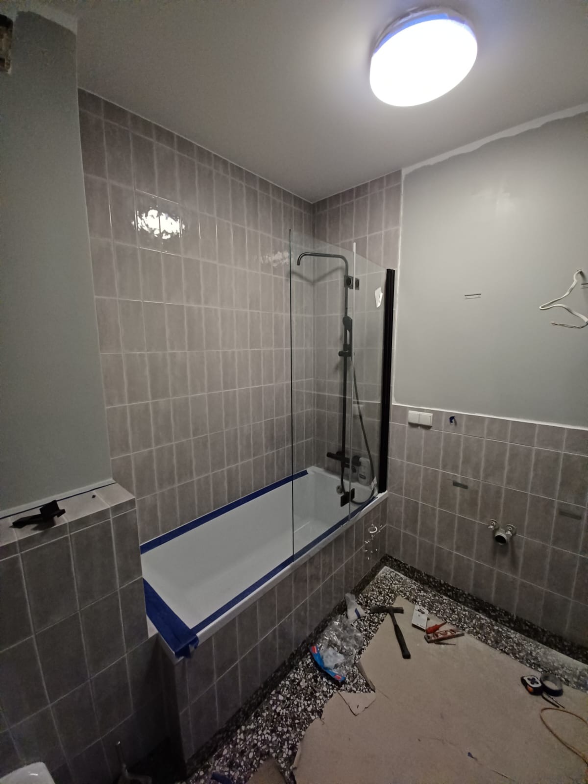 Szara łazienka w trakcie remontu z wanną obudowaną płytkami, szklany parawan nawannowy z czarnymi elementami, widoczne narzędzia i materiały budowlane na podłodze.