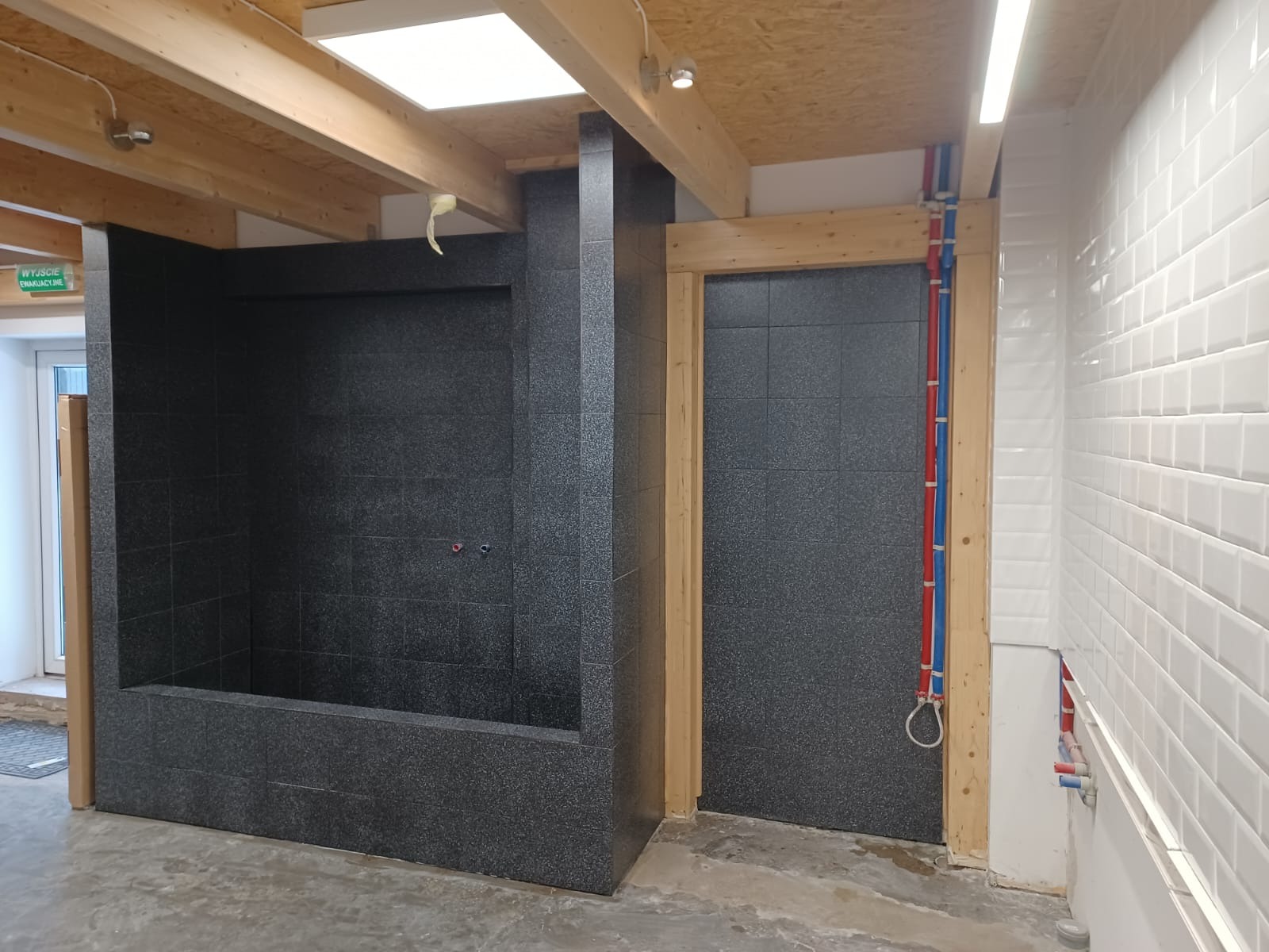 Częściowo wykończona łazienka z czarną mozaiką w strefie prysznicowej i białymi, błyszczącymi płytkami w pozostałej części, widoczne rury instalacyjne w kolorze niebieskim i czerwonym oraz surowy...