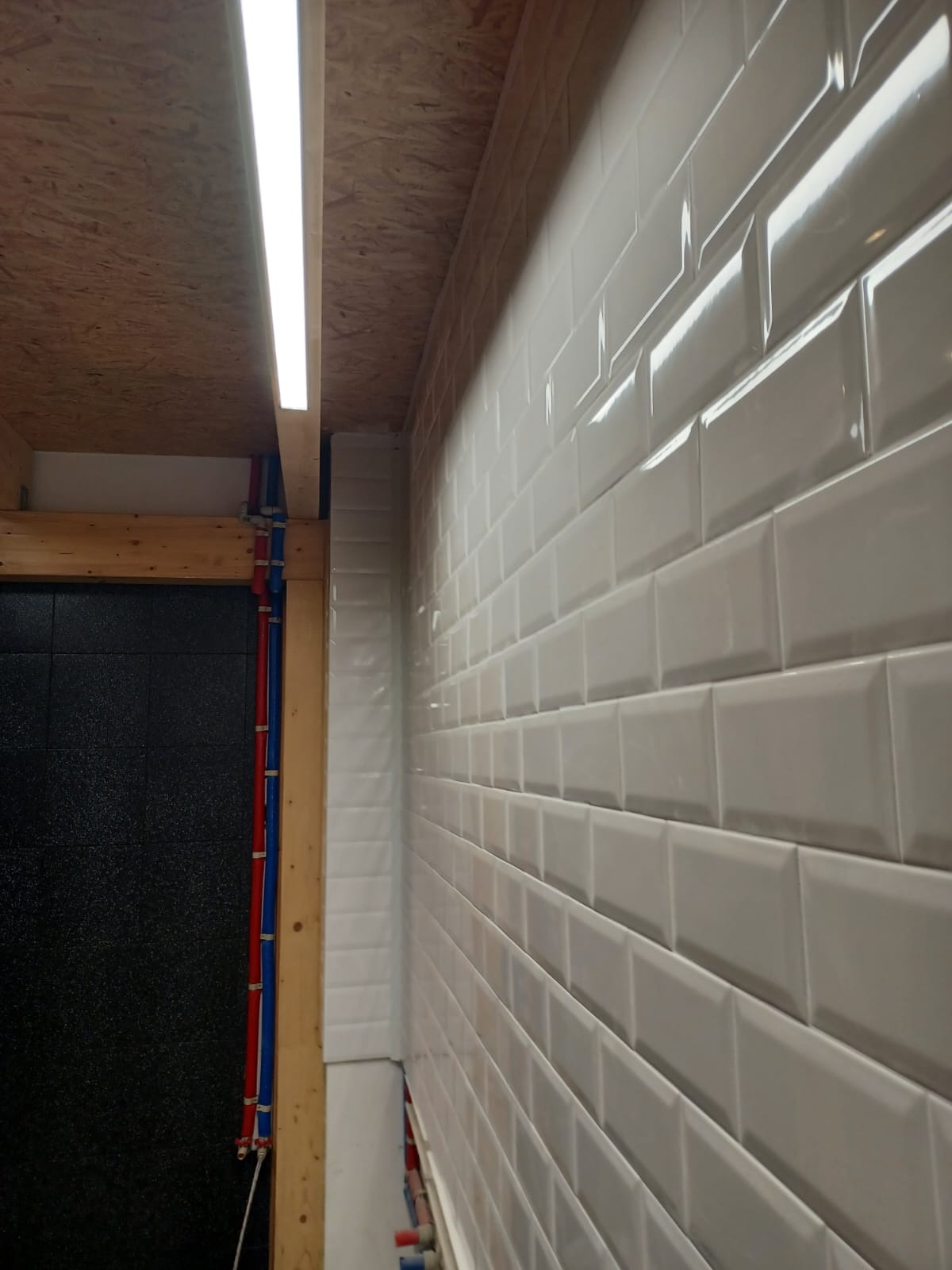 Ściana wyłożona białymi, błyszczącymi płytkami typu metro, widoczne rury instalacji wodnej i surowy strop z płyty OSB z oświetleniem LED w trakcie remontu łazienki.
