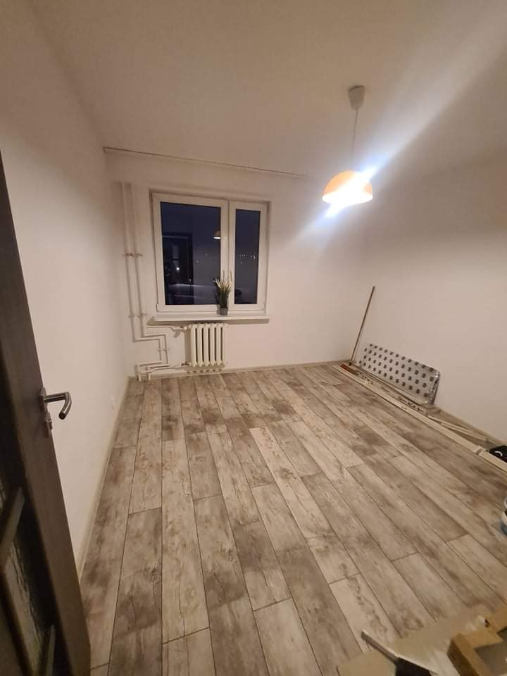 Jasne panele podłogowe imitujące drewno w pustym pokoju z białymi ścianami, widoczne narzędzia i materiały do montażu, okno z grzejnikiem.