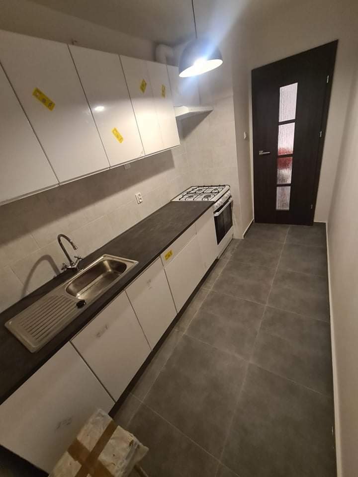 Nowoczesna, wąska kuchnia po remoncie w Warszawie: białe szafki z ciemnym blatem, szare płytki podłogowe, kuchenka gazowa i ciemne drzwi z szybą.