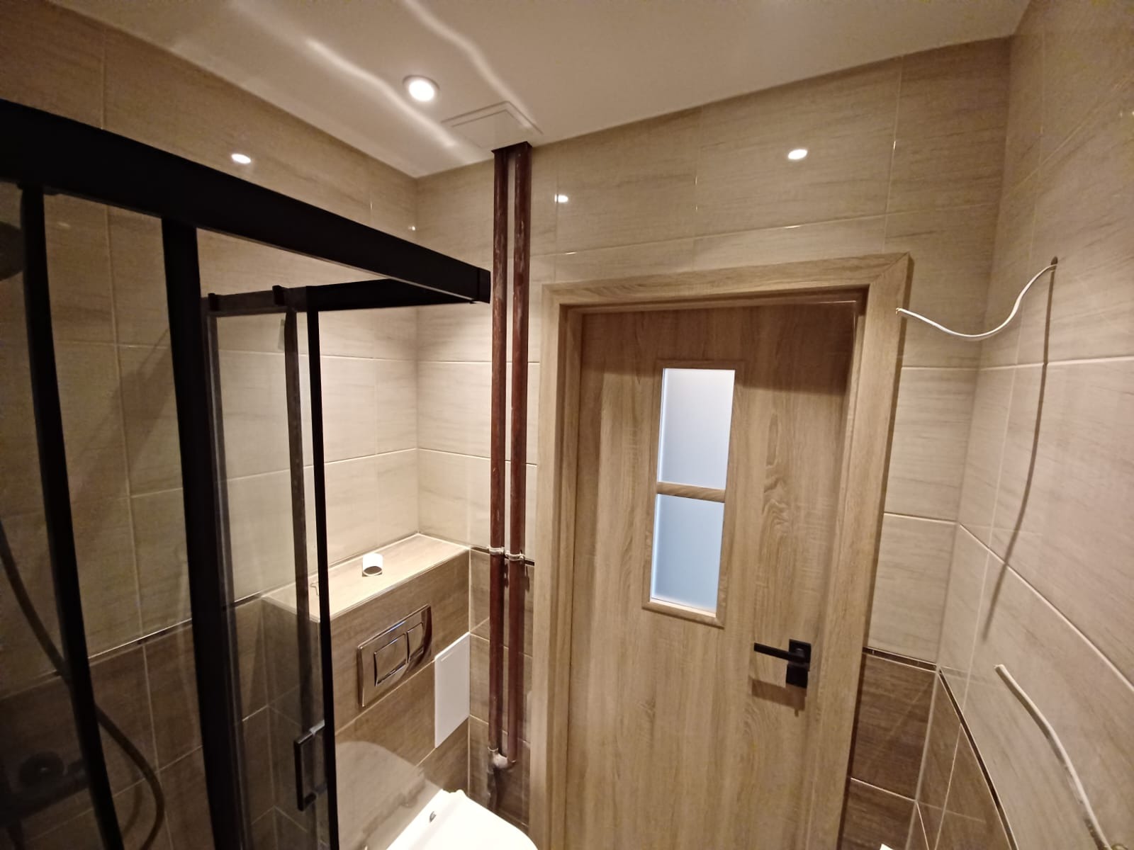 Wykończona łazienka z kabiną prysznicową typu walk-in z czarnymi profilami, beżowymi płytkami imitującymi drewno, drewnianymi drzwiami z szybą i widocznymi miedzianymi rurami.