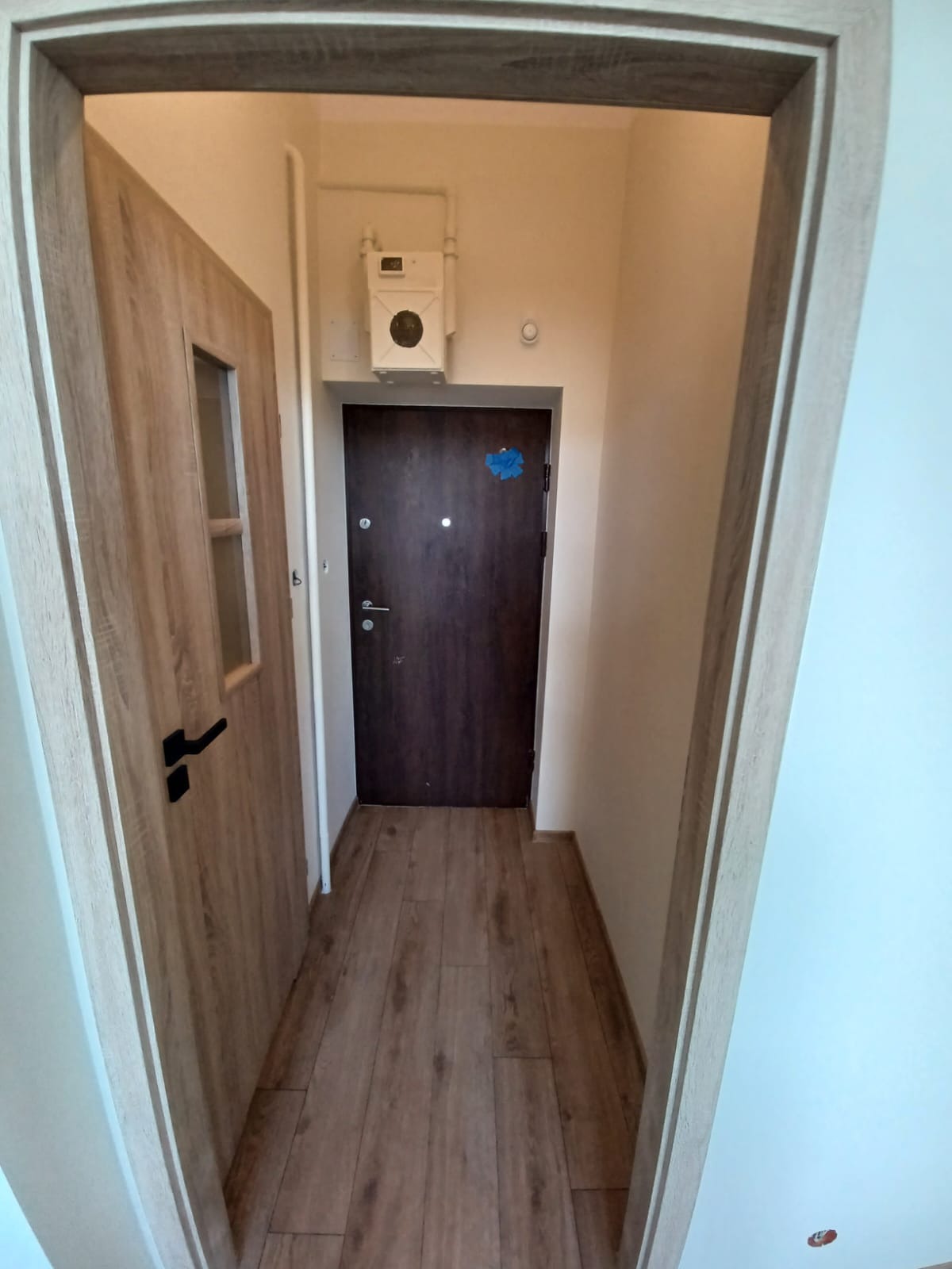 Wnętrze przedpokoju z drewnopodobnymi panelami podłogowymi, dwoje drzwi w drewnianych ościeżnicach, zamek centralny oraz licznik gazu na ścianie.