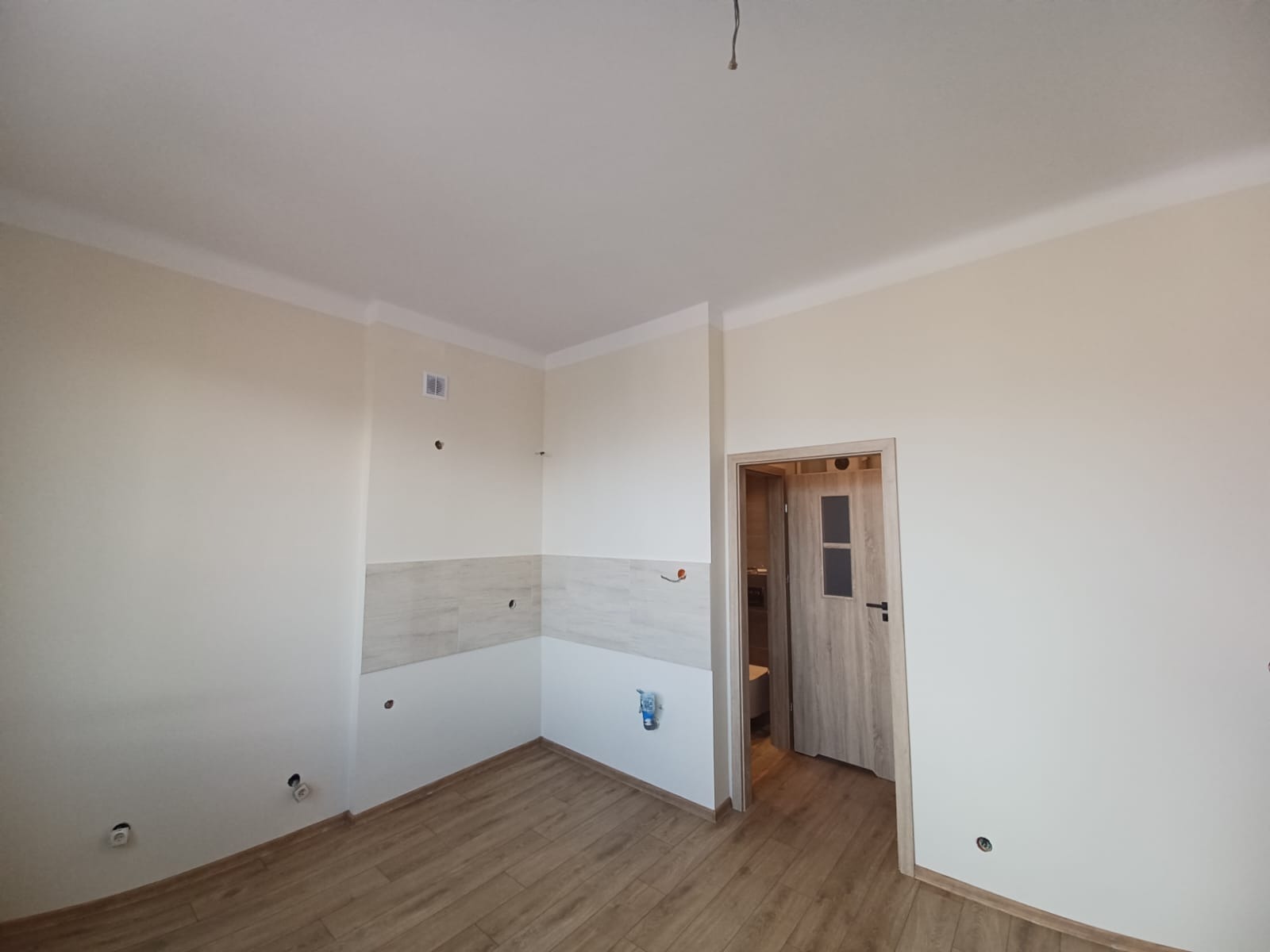 Wnętrze łazienki w trakcie remontu, widoczne częściowo ułożone płytki na ścianie, przygotowane punkty instalacji elektrycznej i hydraulicznej, oraz drewnopodobna podłoga i drzwi wejściowe do łazienki.