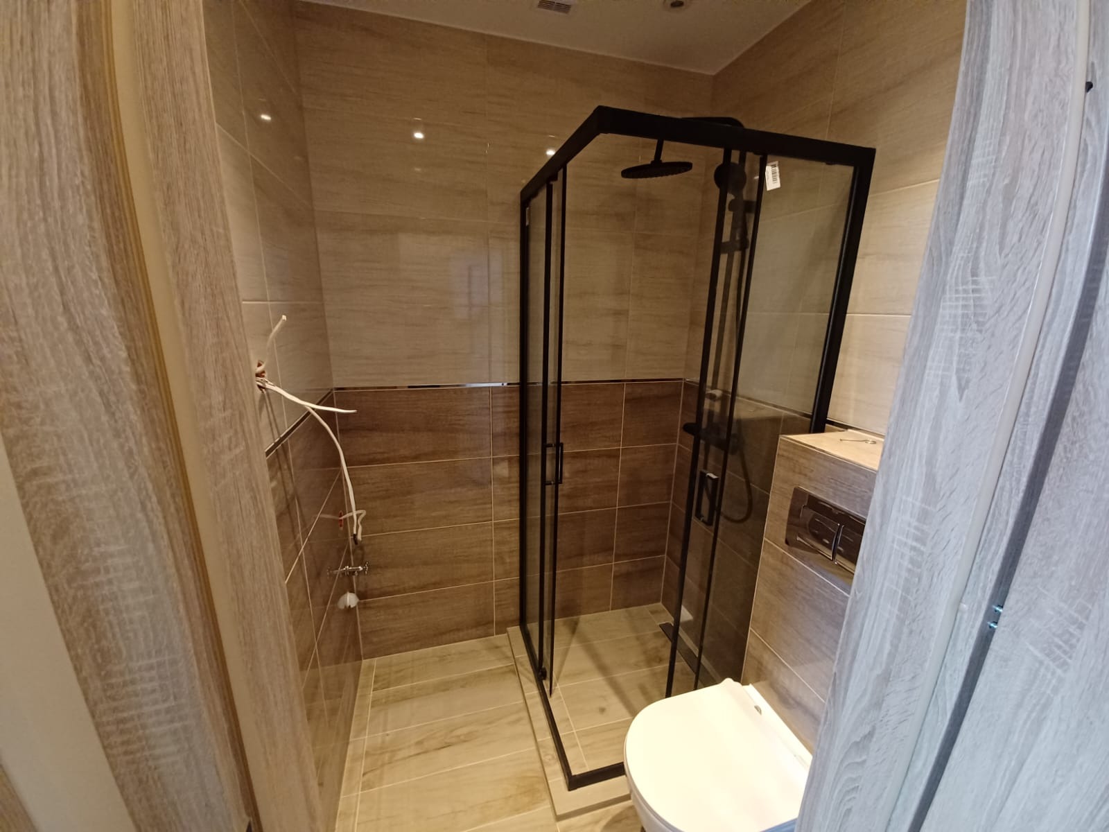Nowoczesna kabina prysznicowa z czarną ramą w łazience wykończonej beżowymi płytkami, widoczny fragment toalety i przyłącza hydrauliczne.