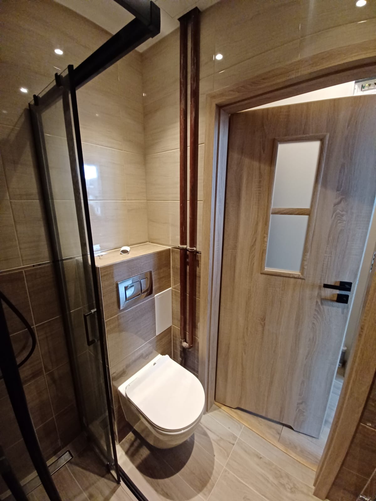 Nowoczesna łazienka z beżowymi płytkami imitującymi drewno, podwieszana toaleta, kabina prysznicowa z czarnymi ramami i drewniane drzwi z przeszkleniem.