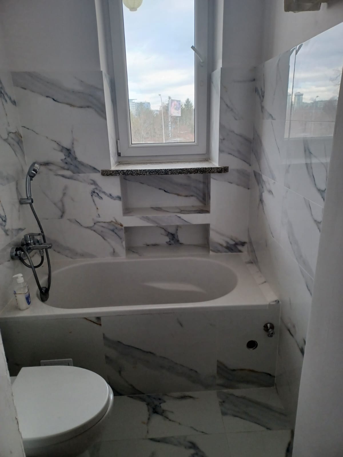 Wykończona łazienka z wanną obudowaną marmurowymi płytkami, z widocznym oknem i wnękami w ścianie.