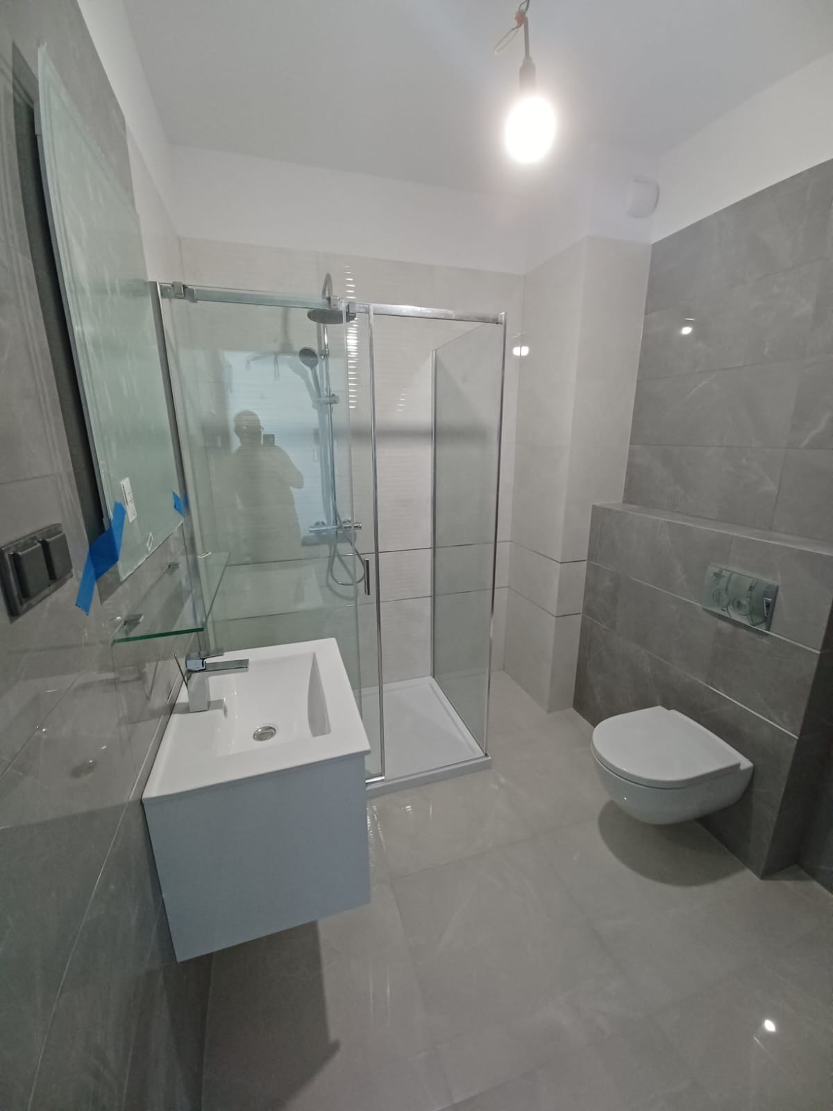 Nowoczesna łazienka w odcieniach szarości z kabiną prysznicową typu walk-in, podwieszaną toaletą i umywalką z szafką, wykończona płytkami imitującymi marmur.