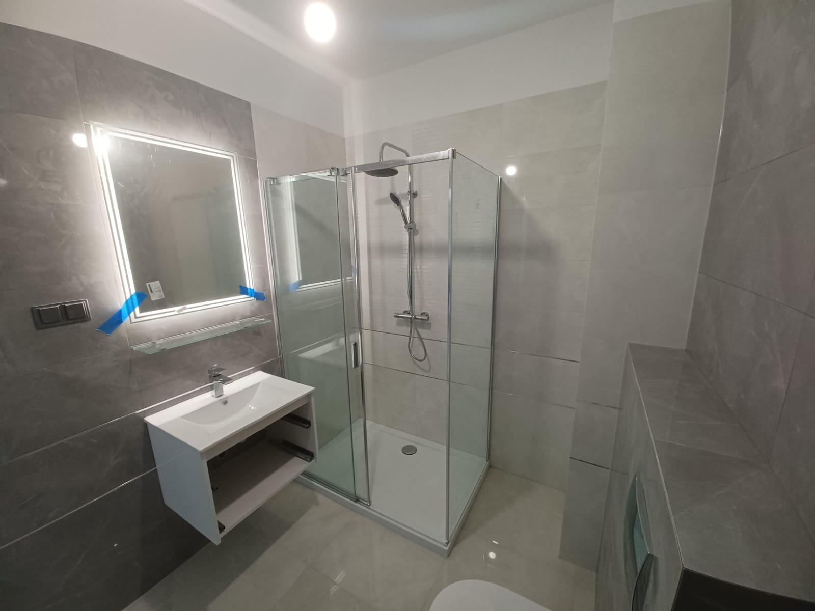 Nowoczesna łazienka z kabiną prysznicową, umywalką z szafką i podświetlanym lustrem, wykończona szarymi płytkami imitującymi marmur, z niebieską taśmą zabezpieczającą na lustrze.