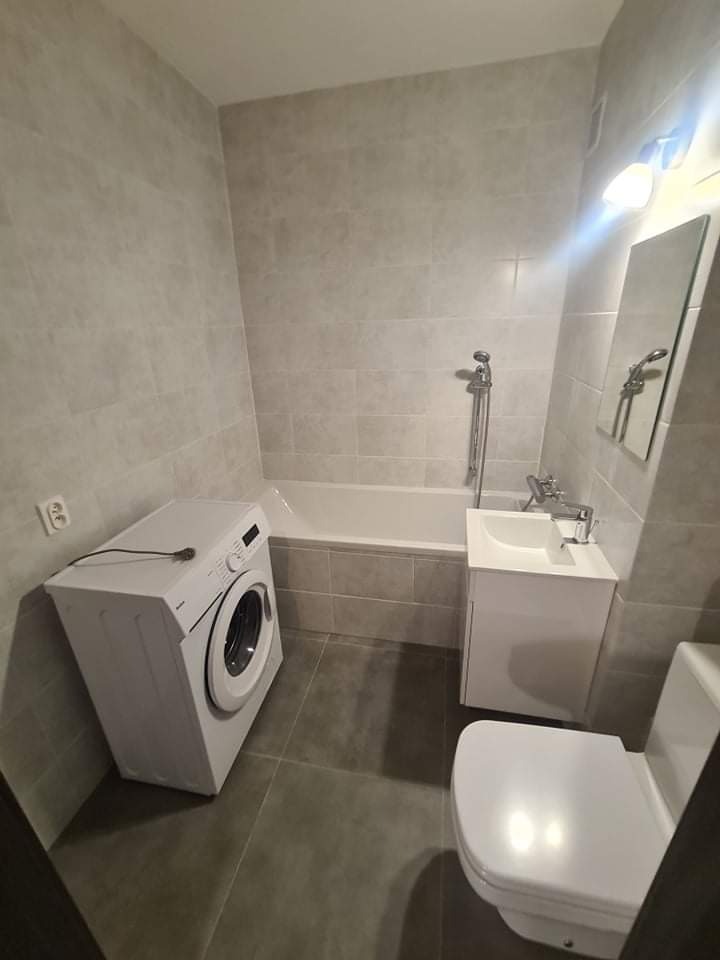 Jasna łazienka po remoncie: szare płytki na ścianach i podłodze, biała wanna, umywalka z szafką i pralka.