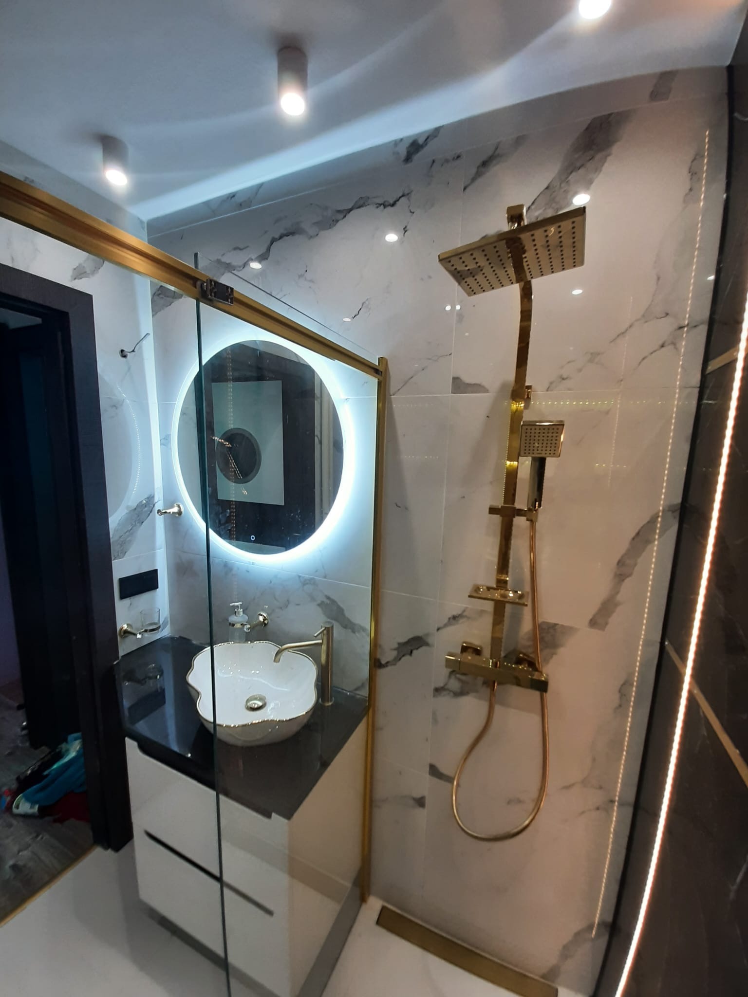 Nowoczesna łazienka z marmurowymi płytkami, złotą armaturą, okrągłym lustrem z podświetleniem LED i umywalką w kształcie kwiatu.