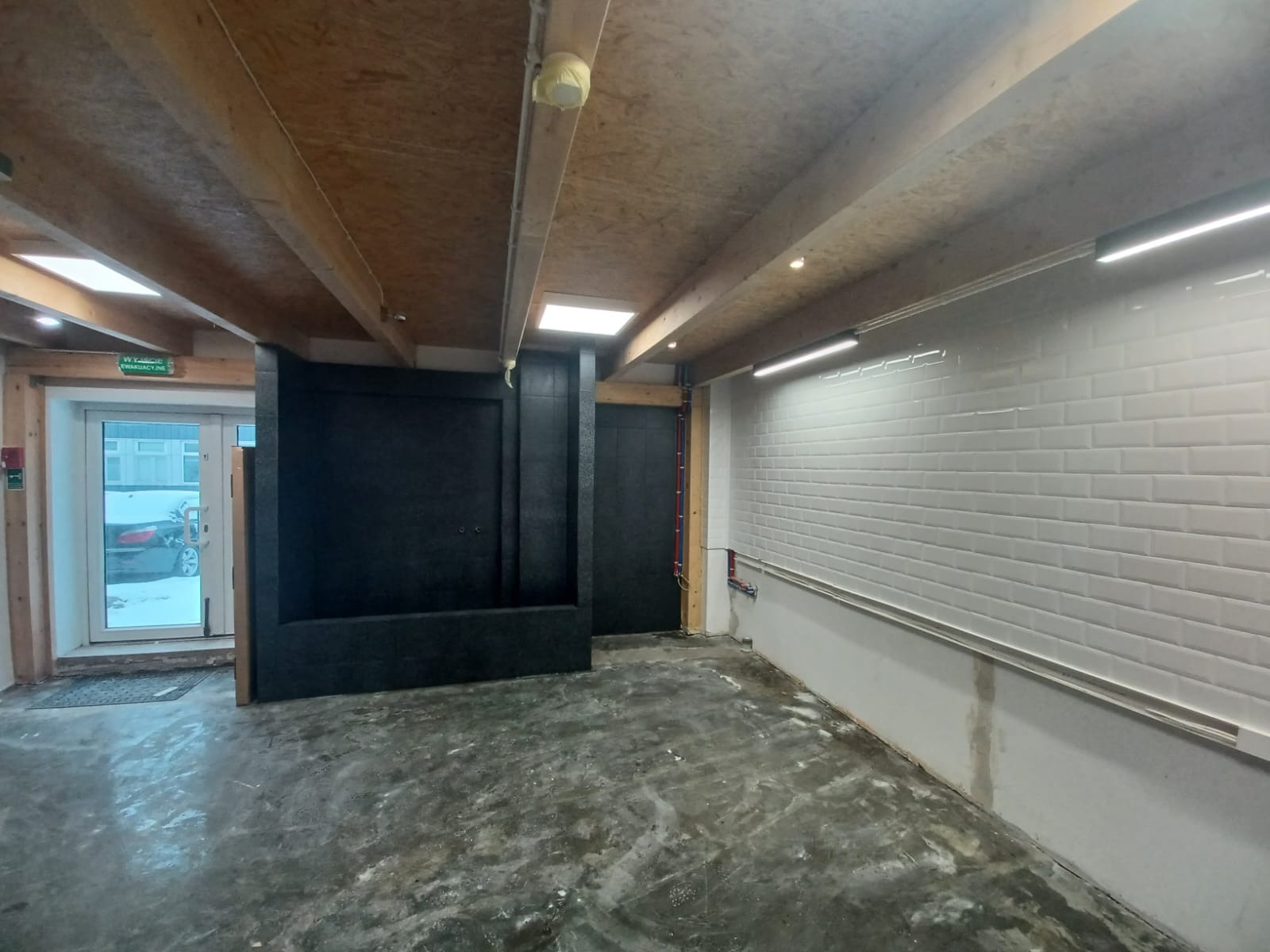 Surowe wnętrze w trakcie remontu: biała ściana z płytek typu metro, surowy beton na podłodze, odsłonięta drewniana konstrukcja sufitu, czarne elementy zabudowy, widoczne instalacje.