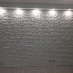 Ściana wyłożona dekoracyjną białą cegłą z widocznymi gniazdkami elektrycznymi, oświetlona punktowymi lampami sufitowymi, na podłodze panele podłogowe.