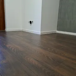 Ułożone ciemne panele podłogowe z widoczną strukturą drewna, białe listwy przypodłogowe i fragment ściany z gniazdkiem elektrycznym.