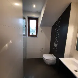 Nowoczesna łazienka z ciemną podłogą, białą ceramiką i czarno-białą ścianą akcentującą, małe okno z rośliną w doniczce, widok na zewnątrz.