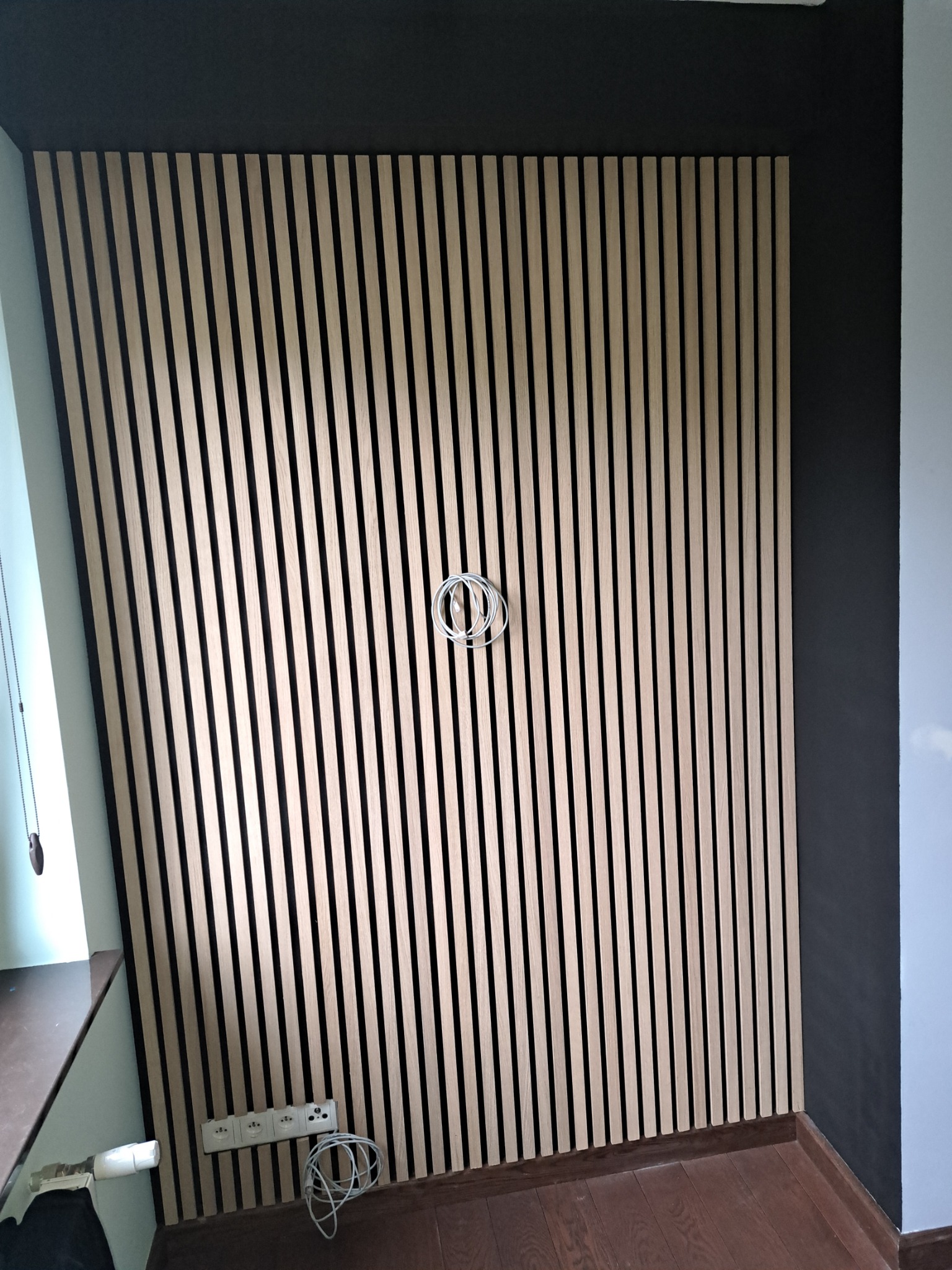 Ściana wykończona panelami lamelowymi w odcieniach beżu i czerni, widoczne gniazdka elektryczne i wystające przewody, sufit pomalowany na ciemny kolor.