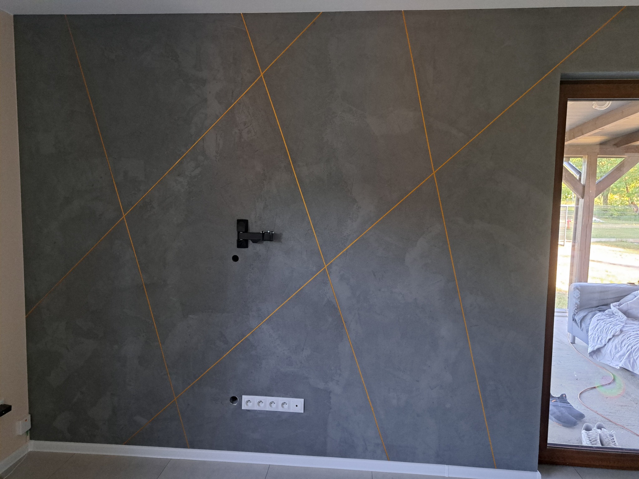 Szara ściana z dekoracyjnymi, złotymi liniami tworzącymi geometryczny wzór, widoczny uchwyt na telewizor i gniazdka elektryczne, fragment okna z widokiem na zewnątrz.